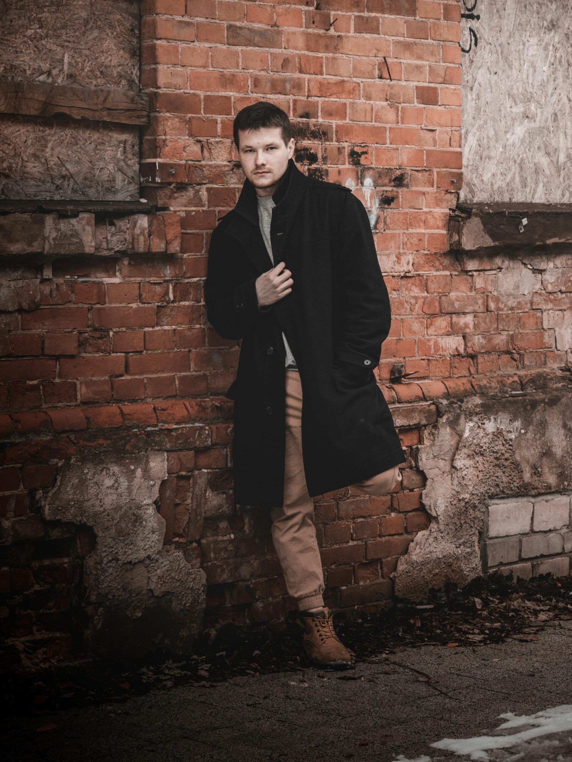 Young man in a black coat leans against a brick wall, showcasing urban fashion style.