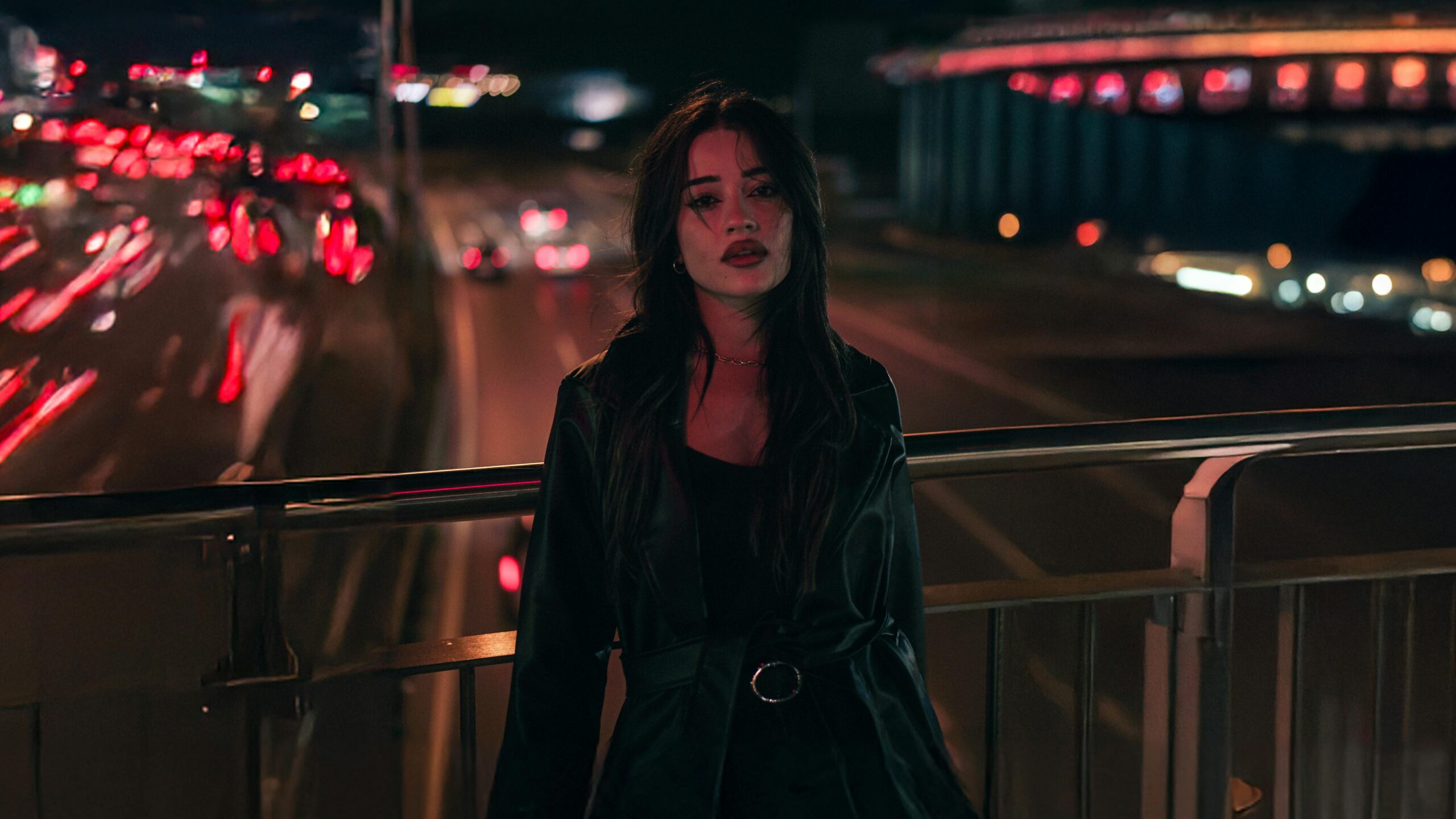 Woman standing on a bridge at night with city lights creating a bokeh effect in the background.