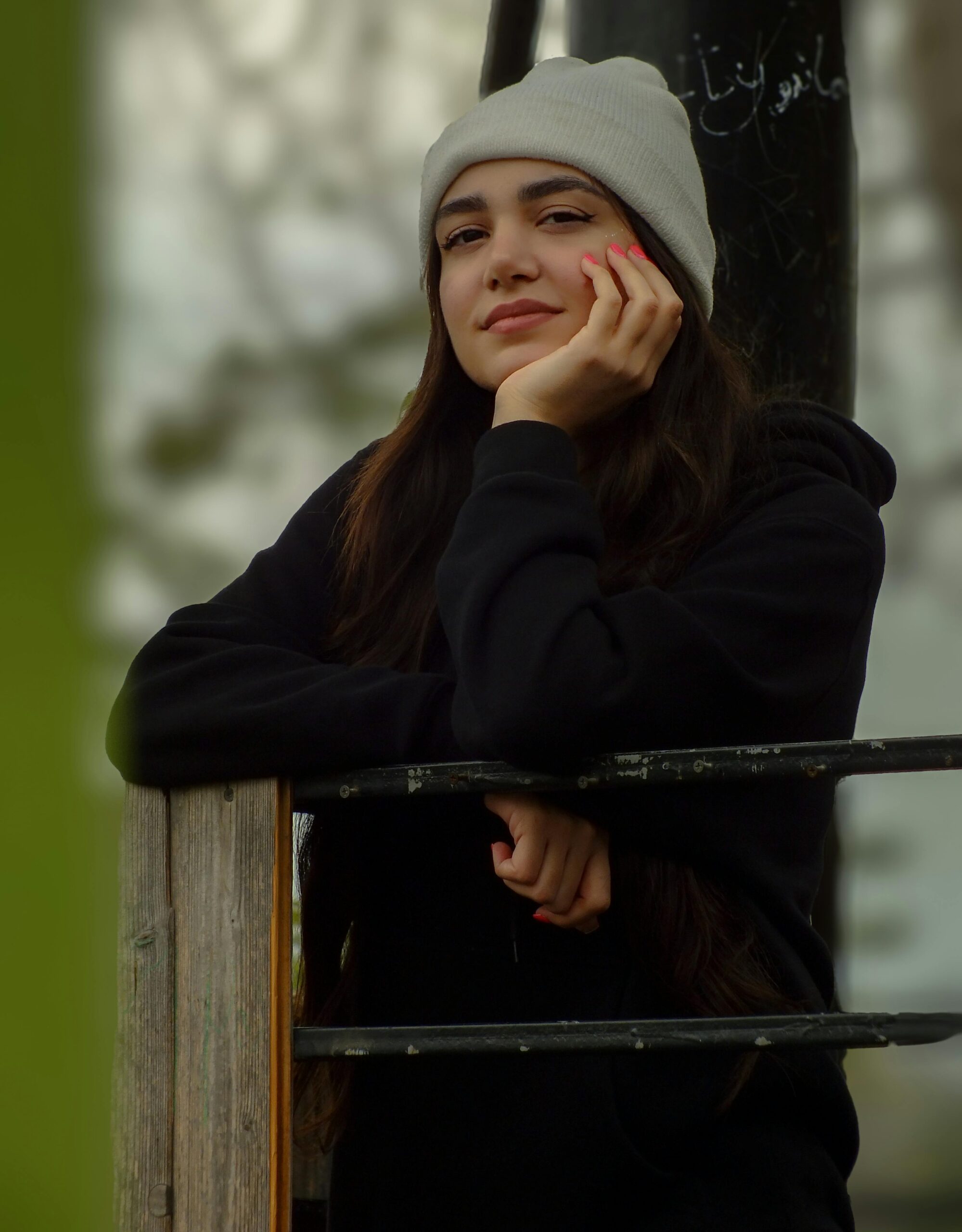 A young woman in a beanie smiles pensively while leaning on a railing outdoors.
