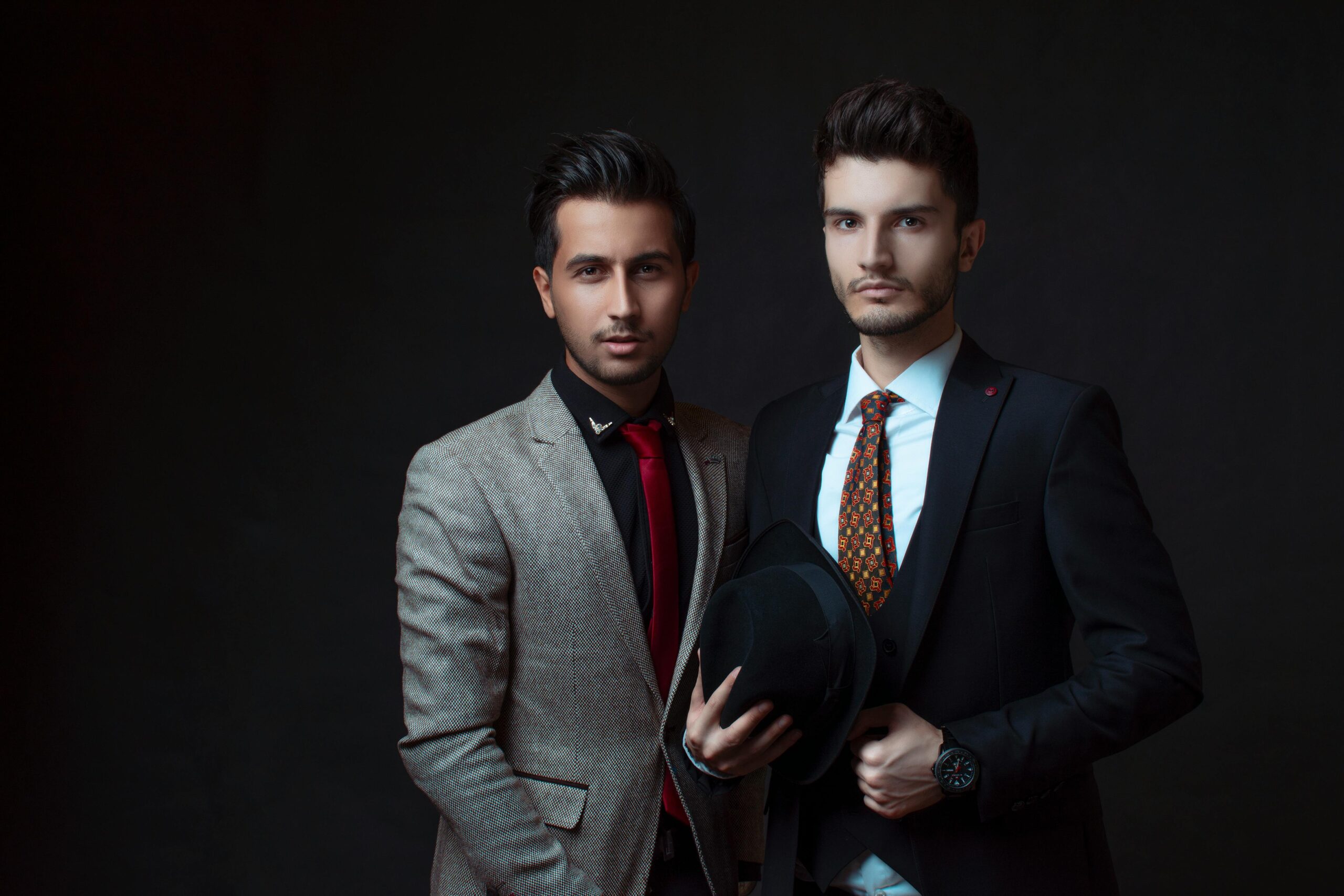 Two men in fashionable suits posing against a dark background, exuding elegance and confidence.
